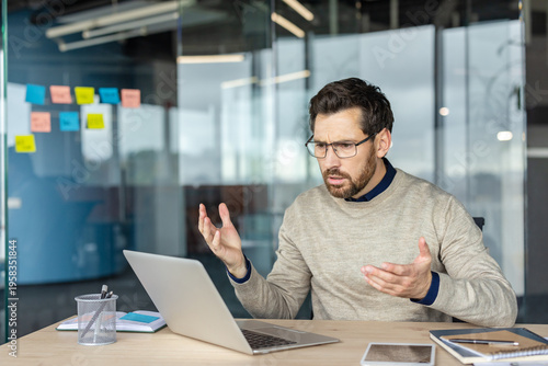 Photos Stressed businessman in glasses sitting at a modern office desk, frustrated and