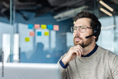 Canvas Print Bearded call center agent with glasses and headset in a modern office, looking a