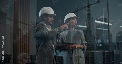Caucasian Male and Asian Female Construction Engineers in Hard Hats Review a Laptop by Window, Focused on Project Finance and Growth. Reflections Reveal Big City Real Estate Buildings and Cranes. 