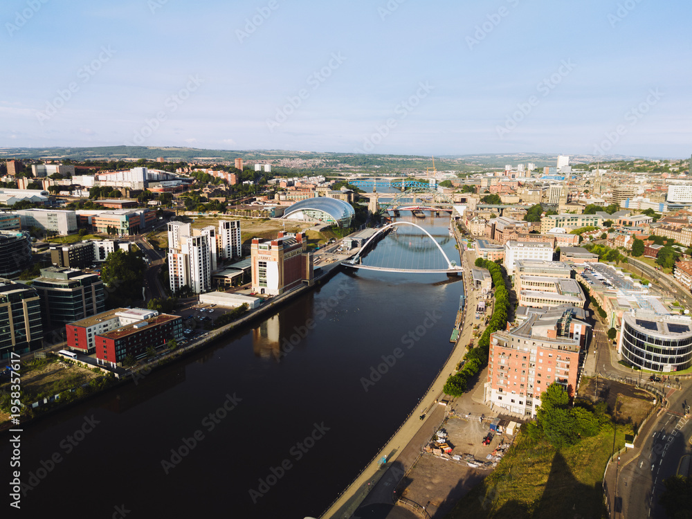 Obraz premium Newcastle upon Tyne: 9th August 2025: Buildings line the Newcastle Quayside at dawn. Bridges stands over the river, reflecting in the water below. Drone view