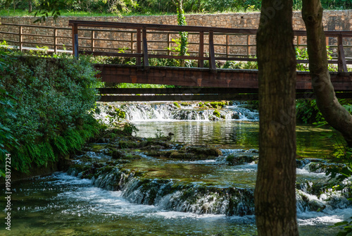 Serene scene of small wooden bridge crossing over gentle, cascading stream surrounded by lush greenery and framed by trees and stone walls. Krka National Park is of Croatia's natural treasures.