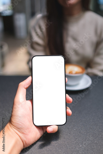 Vertical mockup image of a man holding mobile phone with blank screen with a blurred woman drinking coffee in background