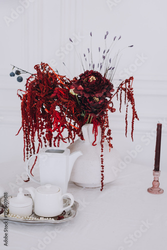Elegant still life with burgundy flowers in a white vase, ceramic tea set, and candle on a white table, ideal for luxury lifestyle and wedding decor backgrounds