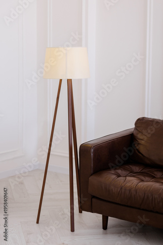 Modern tripod floor lamp with a wooden base and white lampshade standing next to a premium brown leather sofa in a bright minimalist interior, ideal for home decor and furniture design