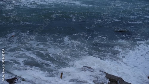 Waves crashing against rocky shore under a cloudy sky. The ocean is turbulent with dark blue water and white foam.