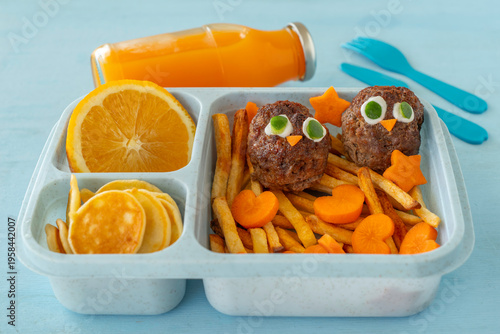 Food background with lunch box of meat balls in the form of chick, fry fries, orange fruit, mini pancakes, carrot, plastic cutlery, bottle of orange juice on blue wood table. Food ideas for kids.
