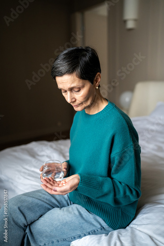 Thoughtful senior woman holding pills and glass of water, unwell elderly female with headache and immune problems taking prescribed supplements to relieve pain, sickness prevention at older age.