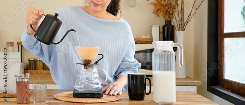 Young asian woman pouring hot water from black kettle into coffee dripper, Female brewing drip coffee in modern kitchen, Morning lifestyle, slow bar cafe and home brewing concept