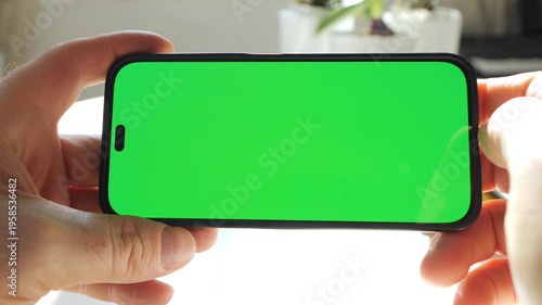 Person holds smartphone with green screen. Fingers tap screen. Background shows indoor plants. Light shines from right side