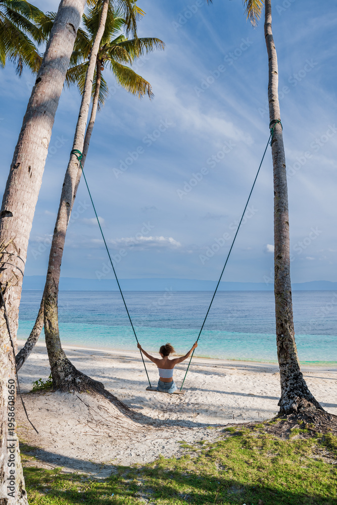 Fototapeta premium Woman on swing between palm trees on beach with ocean view