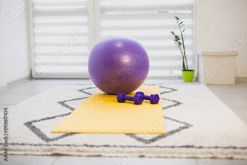 Home gym setup with large purple fitness ball, two dumbbells, and a yellow yoga mat. Home training, healthy lifestyle.