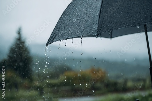 Close-up photo of an umbrella with raindrops falling down, blurry background of greenery and sky in the background, heavy rain.