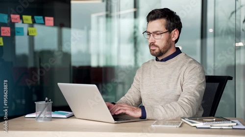 Focused mature businessman working on laptop at desk in office, wearing casual sweater and glasses. Professional typing on computer, managing business tasks, communication and planning in workplace.
