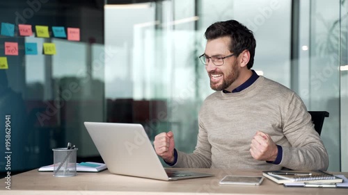 Excited mature businessman reacting to great news on laptop at desk in modern office. Smiling professional celebrates success, feeling surprised, happy while reading message on computer in workplace