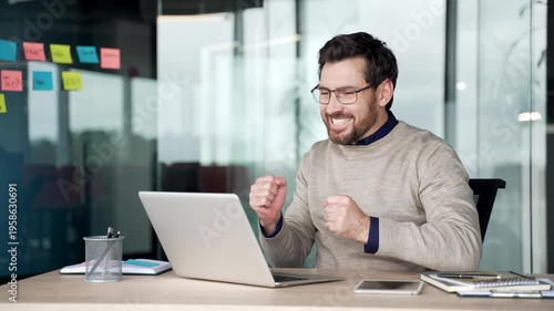 Excited businessman watching online auction on laptop at desk in modern office. Focused mature professional reacts with joy after winning bid, celebrating success and showing enthusiasm at workplace.