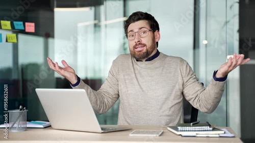 Confused businessman working on laptop at desk in office, then shrugging shoulders and spreading hands while looking at camera. Uncertain mature professional showing doubt and hesitation at workplace.
