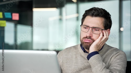 Bored businessman working on laptop, close-up view. Tired mature professional leans head on hand while looking at computer screen, feeling unmotivated and exhausted during routine office work.