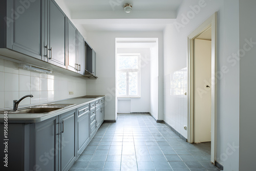An empty kitchen featuring grey cabinets, a sink, and tiled walls/floors. A window and open doorway provide natural light