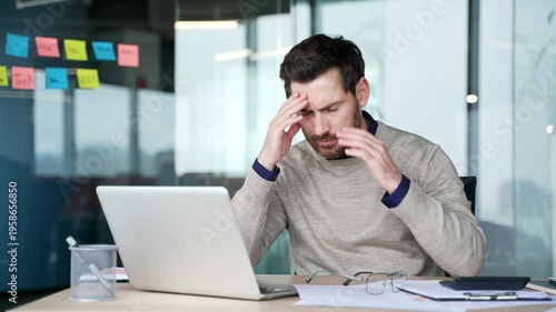 Exhausted businessman working on laptop and paperwork at desk in modern office. Frustrated mature professional facing difficulties, feeling despair and disappointment during stressful work process.