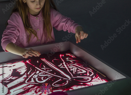 Beautiful little girl draws with sand on a light square, art therapy, sand animation, creative leisure for children, selective focus	