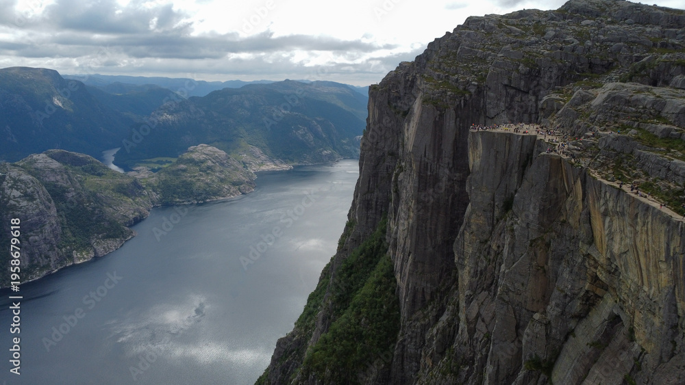 Obraz premium A fjord in Norway is surrounded by steep green slopes, with tourists walking along a winding path high above the deep blue water. Travel brochures for Scandinavian destinations, adventure tourism