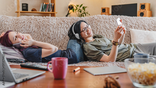 Two girlfriends lying on the sofa at home, enjoying music with headphones and scrolling through their mobile phones, embracing relaxation, digital connection, and cozy moments together.