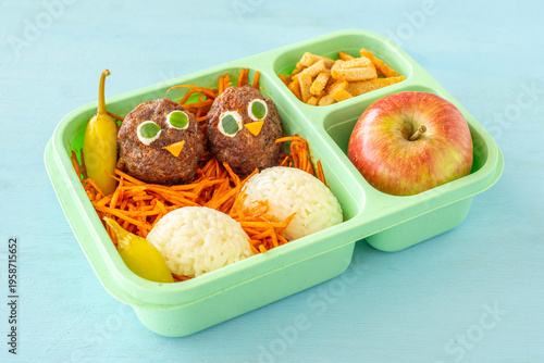 Food ideas for kids. School lunch box with rice, meat balls in the form of chick, fresh carrot salad, red apple.
