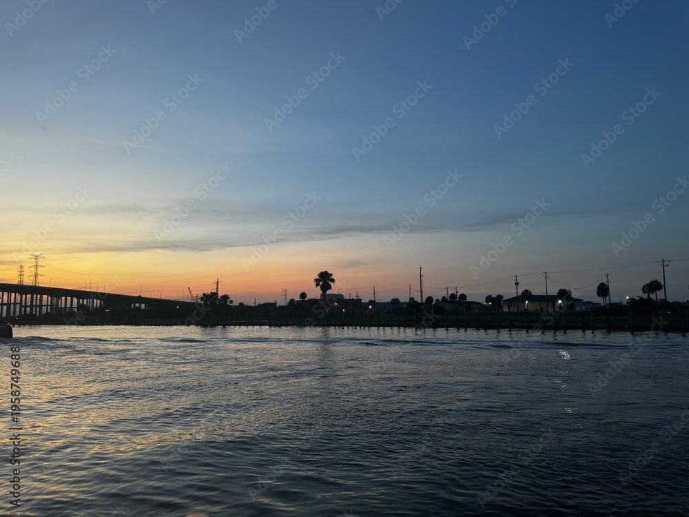 Fototapeta premium Calm water at sunset with a bridge, palm trees, and shoreline structures silhouetted against a soft gradient sky.