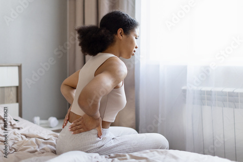 Photos Young african american woman sitting on bed holding lower back in pain, experien