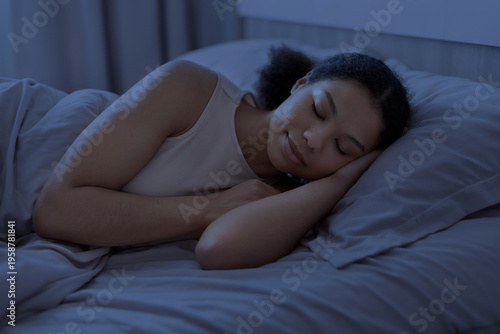 Young black woman sleeping peacefully in bed at night under blanket in cozy bedroom, representing rest, recovery, healthy sleep and calm domestic lifestyle environment