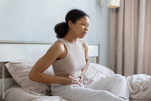 Young black woman sitting on bed holding lower abdomen in pain, showing discomfort and health issues. Domestic bedroom setting highlighting medical and wellbeing concerns