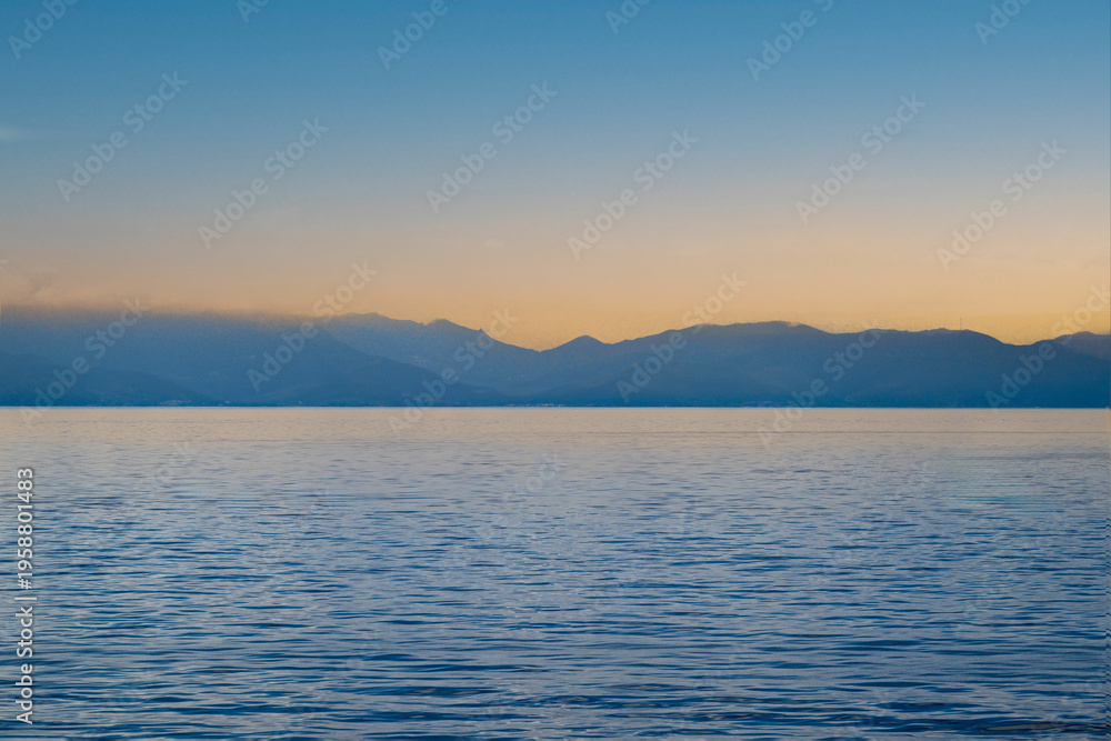 Obraz premium Mer calme bleue avec une chaîne de montagnes lointaine sous un ciel de coucher de soleil pastel au large de l'île de Capraia, Italie