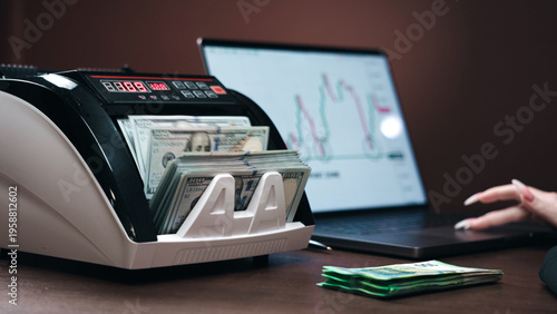 Currency counting machine with stacks of cash and a laptop displaying financial graphs on a wooden desk in a modern office setting