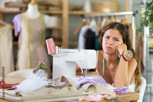Obraz na plátne Young female dressmaker sitting at worktable in front of sewing machine with unf