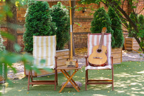 Cozy outdoor relaxation space with wooden garden chairs, acoustic guitar and vintage radio placed on small table. Peaceful backyard leisure scene for music, rest and summer recreation in green garden.