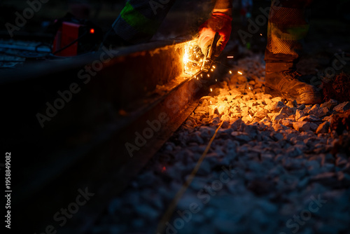 Foto A worker in protective gear grinds a steel rail at night, bright orange sparks fly over crushed ballast stones, calloused hands, glowing cutting wheel, and heavy boots