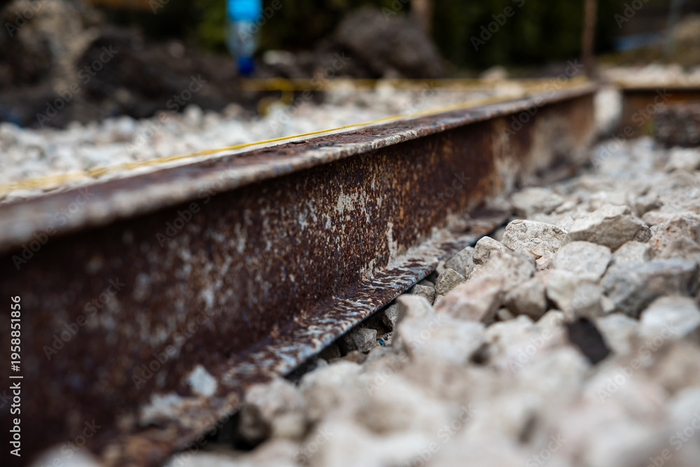 Fototapeta premium A rusted steel rail sits in coarse ballast, pitted and flaked. Shallow depth of field keeps stones sharp and distance soft, daylight tones suggest an outdoor work zone.