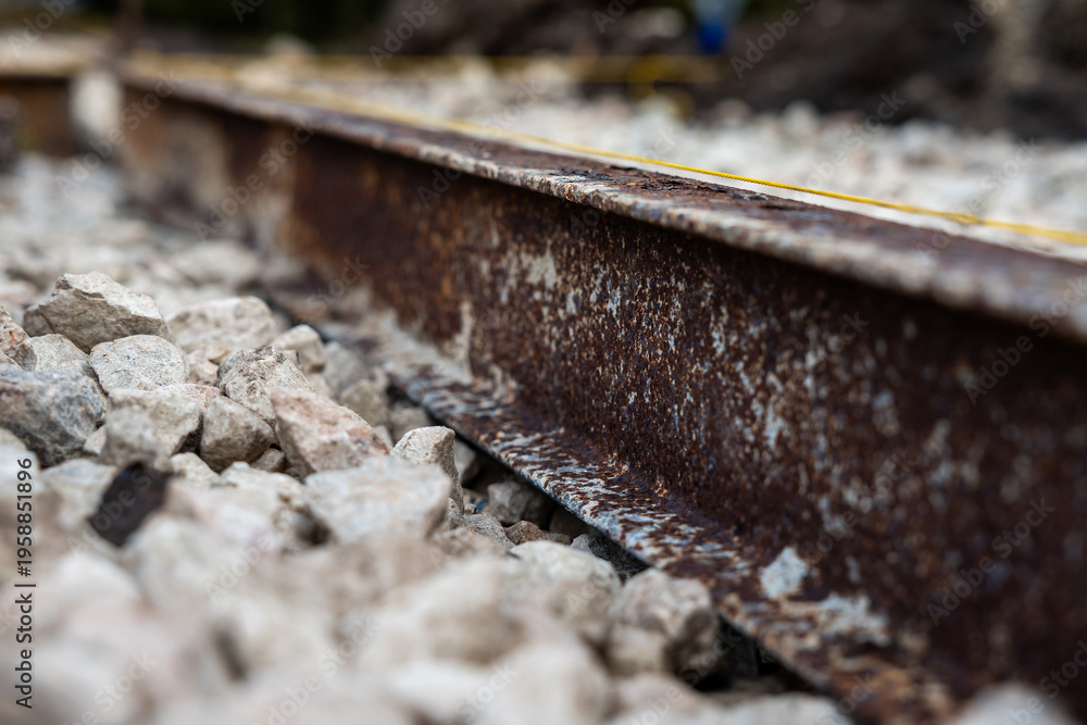 Fototapeta premium Ground level view shows a corroded steel rail, pale angular ballast, and a thin safety line. Late daylight, shallow depth of field, and muted tones emphasize decay.