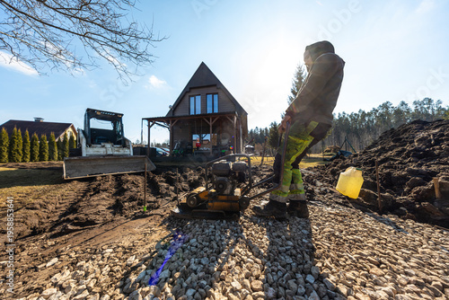 Bilde på lerret A worker in high vis pants runs a plate compactor on fresh gravel as a compact track loader and soil mounds sit before an A frame wooden house in late afternoon light