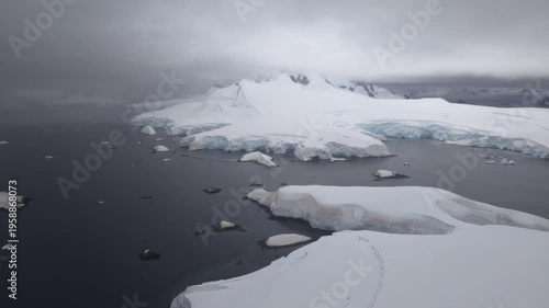 Cinematic aerial moving straight and level over the dark waters of Foyn Harbour, revealing snow-covered volcanic islets and massive stratified ice shelves on the Antarctic Peninsula