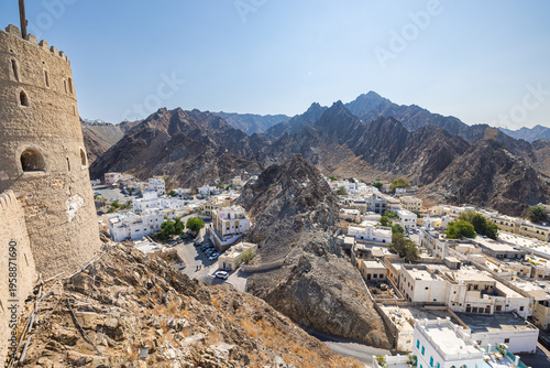 Mountain landscape with traditional white houses in Muscat, Oman, viewed from historic fort walls. Dramatic rocky terrain meets urban settlement under clear blue sky, showcasing Middle Eastern archite