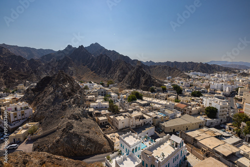 Mountain landscape with traditional white houses in Muscat, Oman, viewed from historic fort walls. Dramatic rocky terrain meets urban settlement under clear blue sky, showcasing Middle Eastern archite