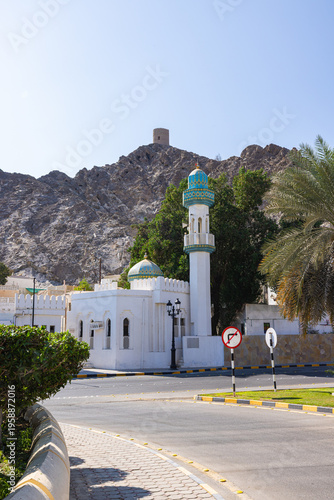 Muscat, Oman – December 28, 2025: Traditional mosque with a turquoise dome and slender minaret located in a quiet residential area of Muscat, set against rugged rocky mountains under a clear blue sky.