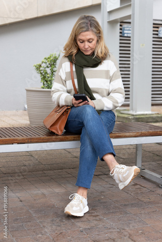 Canvastavla Middle-aged woman sitting on wooden bench in plaza, using smartphone, holding br