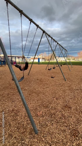 Child on swing at an empty playground during the day under overcast sky. In vertical format.