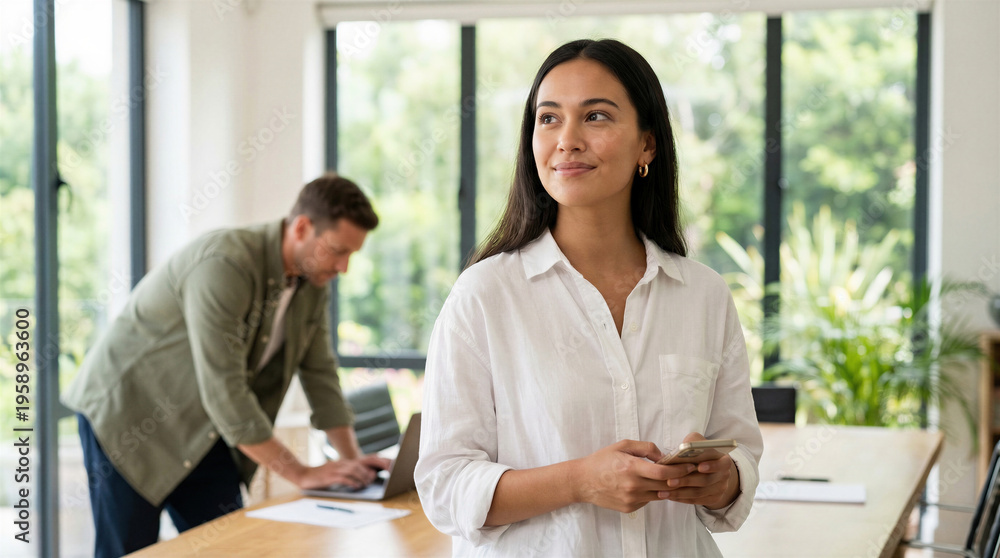 Obraz premium Woman in white shirt standing in office with coworker in background working on laptop