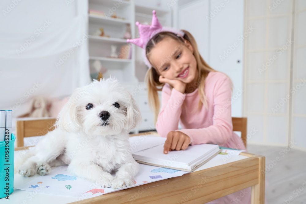 Obraz premium Cute little happy girl with Maltese dog reading book at table in children's bedroom
