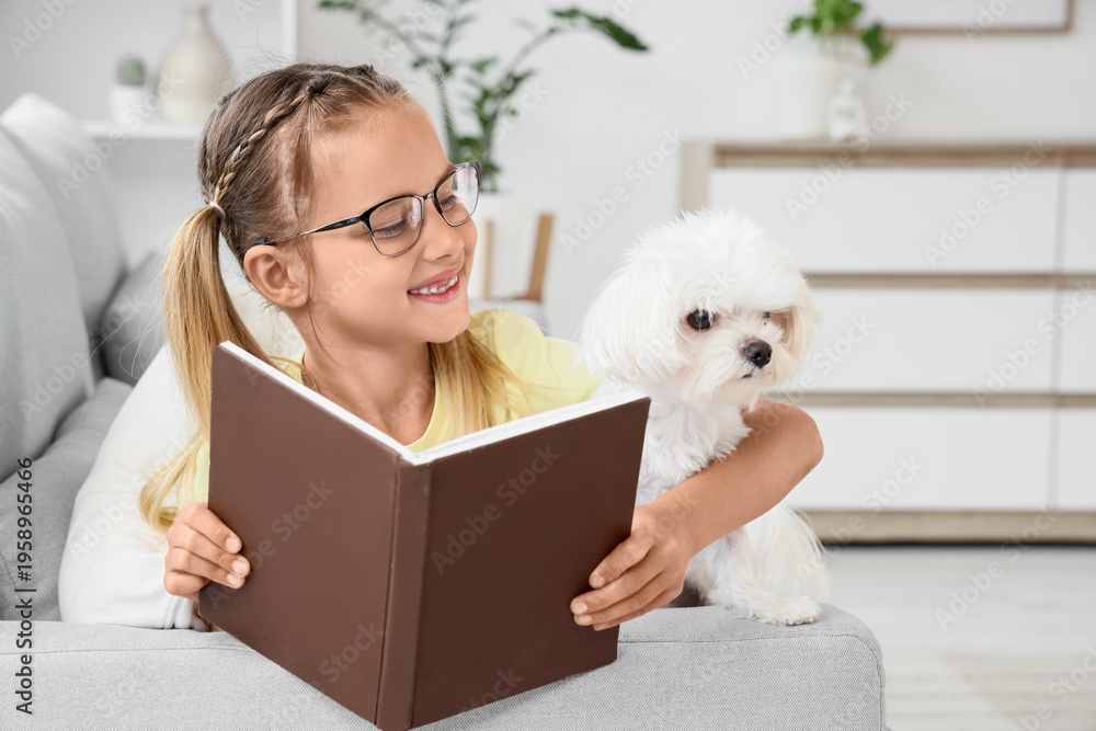 Obraz premium Cute little happy girl with Maltese dog reading book on sofa near window at home
