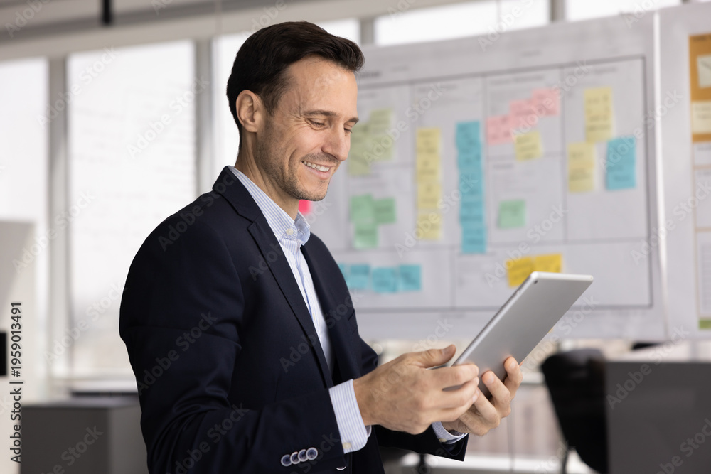 Fototapeta premium Digital planning and analysis. Smiling modern mid aged businessman wearing formal suit use electronic pad device app in work office near agile task board review analytics online optimize project tasks