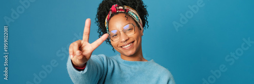 Obraz na płótnie Smiling young woman with glasses dressed in blue sweater shows victory gesture and looks at camera on blue background in studio